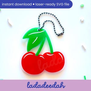 Peut inclure: Un porte-clés en forme de cerise rouge et verte avec une chaîne noire. Les cerises sont soulignées de blanc et les feuilles sont vertes. Le porte-clés est étiqueté "ladadeedah".