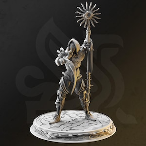 Può includere: Una statuina dettagliata stampata in 3D di un guerriero incappucciato, che tiene in mano un bastone sormontato da un sole a forma di occhio. La figura è vestita con un'armatura ornata e si trova su una base circolare crepata. La tavolozza dei colori è principalmente in tonalità di grigio.