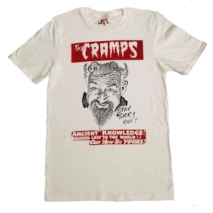 May include: White t-shirt featuring a black and red graphic. The design includes the text "THE CRAMPS" in a red rectangle, a black and white illustration of a bearded man, and the phrases "STAY SICK! KNIF!" and "ANCIENT KNOWLEDGE! BELIEVED LOST TO THE WORLD!! Can Now Be YOURS!" in red.