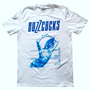 May include: White t-shirt with a blue print of a woman with a cassette tape on her head. The text "BUZZCOCKS" is printed in blue at the top of the shirt. The text "DICT AD ASM OIRGA" is printed vertically on the right side of the shirt.