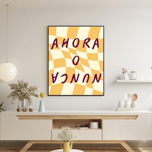Könnte beinhalten: Ein gerahmter Kunstdruck mit den Worten "AHORA O NUNCA" in Lila auf einem gelb-cremefarbenen Schachbrettmuster. Das Kunstwerk wird auf einem weißen Schrank mit dekorativen Gegenständen ausgestellt.