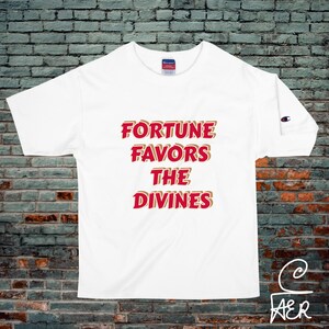 Könnte beinhalten: Weißes T-Shirt mit dem Text "FORTUNE FAVORS THE DIVINES" in roter und goldener Schrift. Das Shirt hat kurze Ärmel und ein Champion-Logo auf dem linken Ärmel.