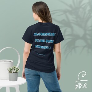 May include: A dark navy blue short-sleeved t-shirt featuring the text ALCHEMIZE YOUR OWN ENERGY! in a bold, light blue font on the back. The shirt is paired with blue denim jeans, creating a casual look with clear, legible lettering on the fabric.