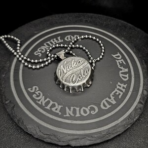 May include: A silver-colored pendant necklace featuring a bottle cap design with the words "Nuka Cola" in a stylized font. The necklace rests on a dark, circular coaster with engraved text. The overall aesthetic is vintage and industrial.