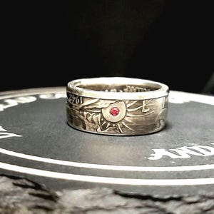 May include: A silver ring made from a modified US half dollar coin with a red gemstone set in the center. The ring is sitting on a black slate coaster with the words "Dead  Heads and  Trucks" inscribed on it.