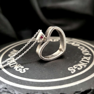 May include: A silver heart-shaped pendant necklace with a small red gemstone. The pendant has a textured, ridged design. The necklace is displayed on a dark, textured surface with the words "CAP COIN RINGS".