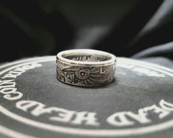 May include: Silver ring with a detailed design featuring a red stone and the number 1961. The ring is on a black surface with the words "Deadwood" and "Company" visible.