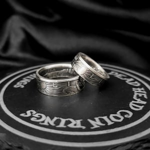 May include: Two silver coin rings with detailed designs, one slightly smaller than the other, rest on a black surface. The rings feature intricate engravings of sun and wave patterns. The background is a dark, textured fabric.
