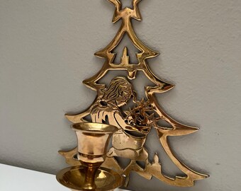May include: A gold-coloured Christmas tree-shaped wall sconce with a star on top. The sconce features a detailed cutout design of a person holding a basket. The sconce has a candle holder with a small dish.