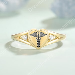 May include: A gold ring featuring a heart-shaped emblem with a black caduceus symbol, flanked by two small, clear gemstones. The ring has a delicate band and a polished finish, suitable for medical professionals or those in healthcare.