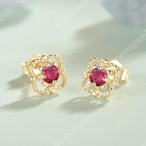 May include: A pair of gold-colored stud earrings. Each earring features a round, bright red gemstone set in a four-leaf clover design. The earrings are displayed on a light green surface.