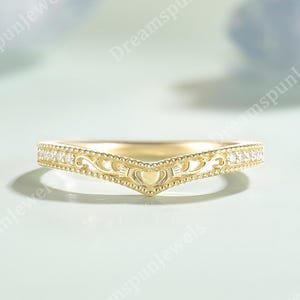 May include: A gold wedding band with a V-shaped design. The ring features a heart-shaped centre and is adorned with small, clear gemstones and intricate filigree detailing. The ring is set against a soft, light blue background.
