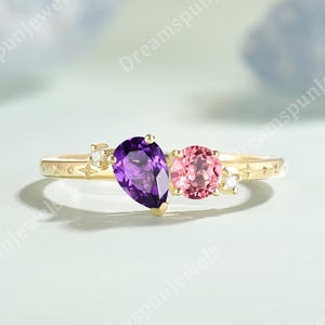 May include: A gold ring featuring a pear-shaped purple amethyst and a round pink tourmaline gemstone. The band is accented with small clear stones and engraved star and cross symbols along the metal surface, creating a detailed jewellery piece.