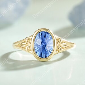 May include: A gold ring with an oval, blue gemstone. The ring has leaf detailing on the shoulders. The gemstone is set in a bezel setting, and the band is smooth. The ring is a piece of jewellery.