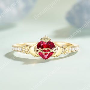 Peut inclure: Une bague Claddagh dorée avec une pierre rouge en forme de cœur au centre. La bague présente une couronne au-dessus du cœur et des mains de chaque côté. De petits gemmes transparents ornent la bande.