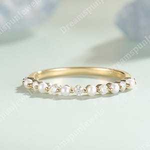May include: A gold band ring with alternating small pearls and clear gemstones. The ring is set against a soft, light green background. The ring is a delicate piece of jewellery.