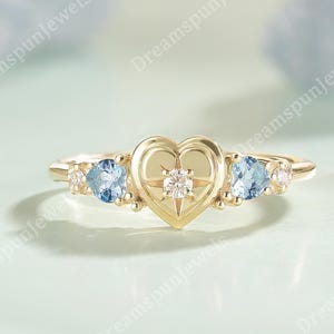 Peut inclure: Une bague en or avec un cœur central orné d'un motif en étoile et d'un petit diamant. Deux pierres bleues claires et de petites pierres d'accentuation flanquent le cœur. La bague a un anneau délicat et est posée sur un fond vert clair et doux.