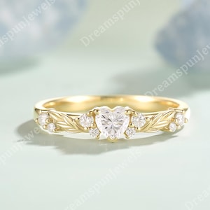 Peut inclure: Une bague en or avec un diamant central en forme de cœur. La bande est ornée de feuilles et de petits diamants ronds. La bague est posée sur un fond bleu clair et doux, mettant en valeur les détails du bijou.