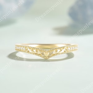 Peut inclure: Une bague dorée en forme de V présentant un motif Claddagh central avec un cœur, des mains et une couronne. L'anneau est orné de petites pierres claires sur les côtés et d'un travail de filigrane complexe, formant une silhouette en chevron.