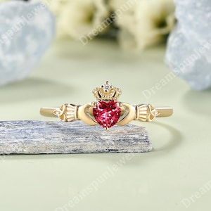 May include: A gold Claddagh ring featuring a central heart-shaped pink gemstone topped with a small crown. The band displays intricate metalwork details on both sides, showcasing a traditional design with a polished finish and a clear, vibrant stone.