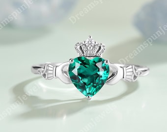 May include: A silver-toned Claddagh ring with a heart-shaped, emerald-green gemstone. The ring features a crown above the heart and hands on either side. The ring is set against a soft, blurred background.