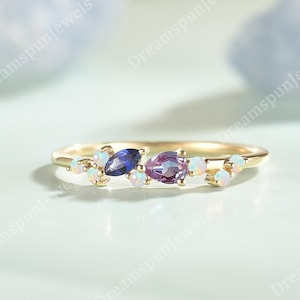 May include: A gold ring with a cluster of gemstones. The ring features a central purple pear-shaped stone, a blue oval stone, and several small white stones. The ring is set against a light blue background.
