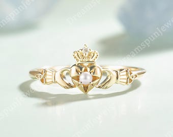 May include: A gold Claddagh ring featuring a central heart shape with a small white pearl at its center, topped with a detailed crown. The band displays intricate metalwork on both sides, extending from the hands that hold the heart.