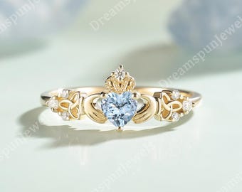 May include: A gold Claddagh ring featuring a central heart-shaped light blue gemstone, a crown detail, and two hands holding the heart. The band is accented with intricate knotwork patterns and small clear round stones on both sides of the center.