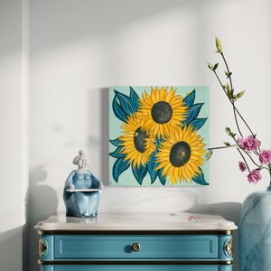 May include: A painting depicting three sunflowers with dark centres and yellow petals set against a light blue background. The sunflowers are surrounded by dark green leaves.