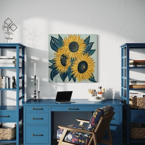 May include: A painting of three sunflowers with dark blue leaves on a light blue background. The sunflowers are yellow with dark brown centres. The painting is hanging on a white wall above a blue desk with a laptop, a chair, and two bookshelves.