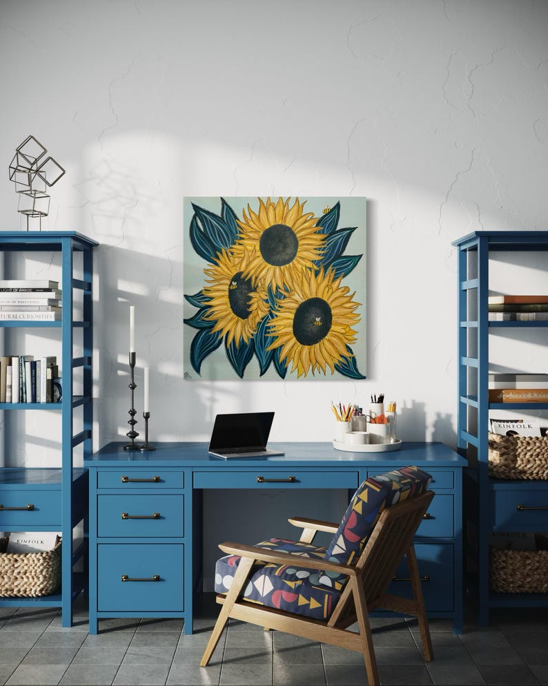 May include: A painting of three sunflowers with dark blue leaves on a light blue background. The sunflowers are yellow with dark brown centres. The painting is hanging on a white wall above a blue desk with a laptop, a chair, and two bookshelves.