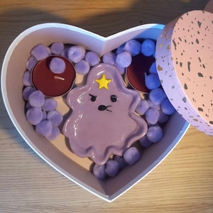May include: A heart-shaped box with a purple Lumpy Space Princess character, two red candles, and purple pom-poms.
