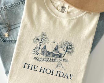 May include: Cream-colored t-shirt with a navy blue illustration of a house in a winter landscape. The text "THE HOLIDAY" is printed below the image. The shirt is paired with light-wash denim jeans and a tan hat.