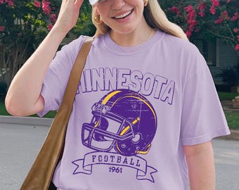 Puede incluir: Una camiseta de color lila claro con la palabra "MINNESOTA" sobre un gráfico de casco de fútbol morado y dorado. Debajo del casco está el texto "FOOTBALL 1961". La persona lleva una gorra de béisbol blanca y pantalones cortos blancos.
