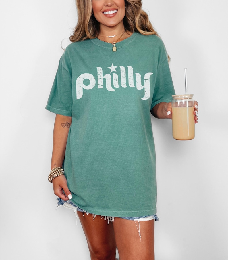 May include: A green t-shirt with the word "Philly" in white, featuring a star above the "y". The shirt is paired with denim shorts. A glass jar with a straw and a beige beverage is held in the hand.