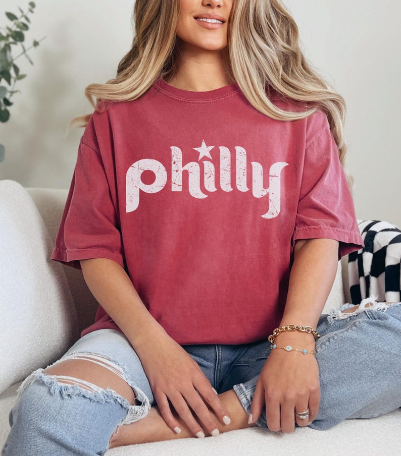 May include: A maroon t-shirt with the word "philly" in white, stylized text. A white star is above the "y". The shirt is worn with blue jeans. The shirt has a vintage, worn look.