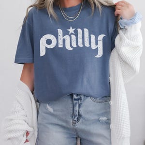 May include: A blue t-shirt with the word "philly" in white, featuring a star above the "y". The shirt is paired with denim shorts and a white cardigan. The overall style is casual and comfortable.