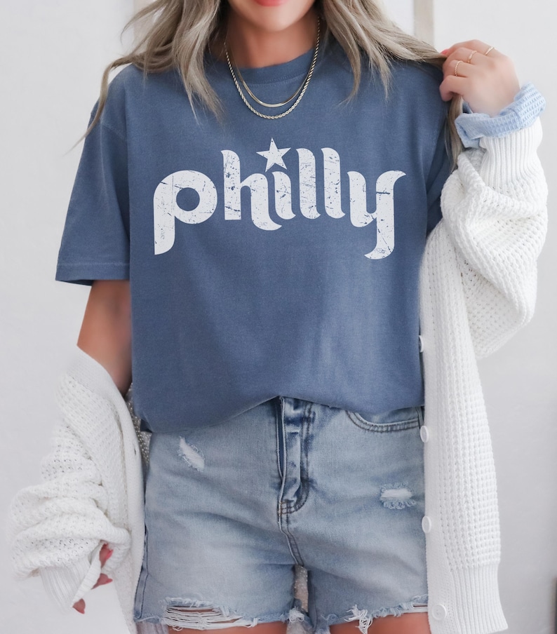 May include: A blue t-shirt with the word "philly" in white, featuring a star above the "y". The shirt is paired with denim shorts and a white cardigan. The overall style is casual and comfortable.