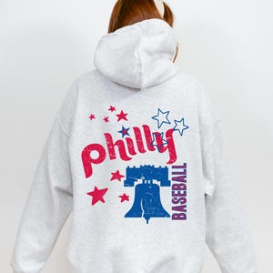 Könnte beinhalten: Grauer Kapuzenpullover mit einem roten und blauen Grafikdruck der Liberty Bell und dem Wort "philly" mit Sternen. Der Text "BASEBALL" ist vertikal auf der rechten Seite des Grafikdrucks gedruckt.