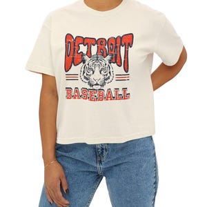 Puede incluir: Una camiseta blanca recortada con un gr&aacute;fico de estilo vintage de una cabeza de tigre y el texto "DETROIT BASEBALL" en rojo y azul.