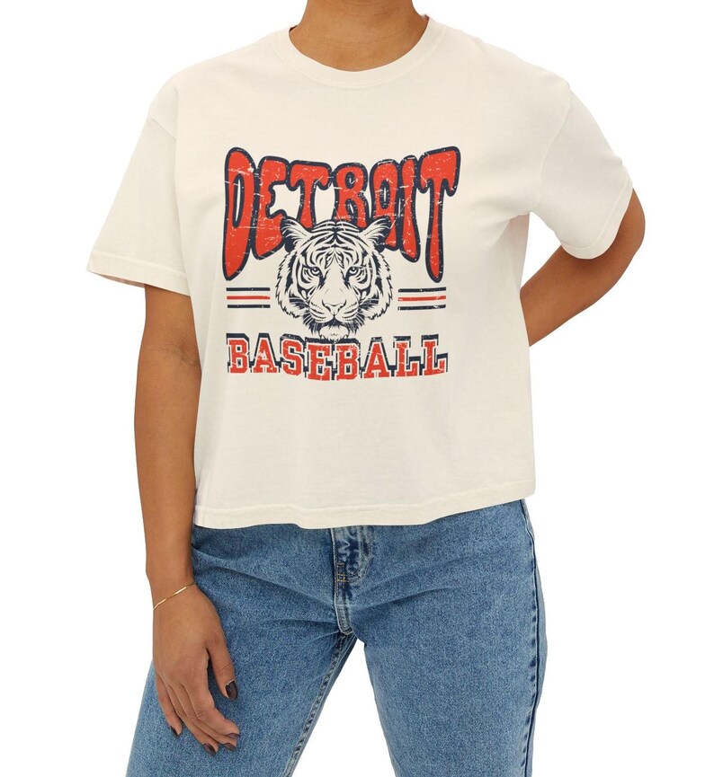 Puede incluir: Una camiseta blanca recortada con un gr&aacute;fico de estilo vintage de una cabeza de tigre y el texto "DETROIT BASEBALL" en rojo y azul.
