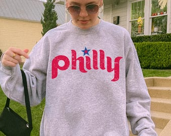 May include: A heather gray crewneck sweatshirt featuring the word philly printed in a distressed red font across the chest, with a small blue star positioned above the letter i. The garment has long sleeves and a ribbed neckline.