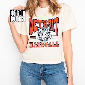 Puede incluir: Una camiseta blanca recortada con un gr&aacute;fico de estilo vintage de un tigre y el texto "DETROIT BASEBALL" en naranja y azul.