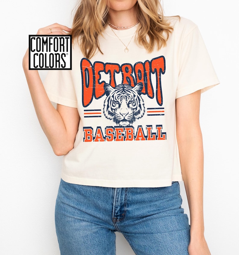 Puede incluir: Una camiseta blanca recortada con un gr&aacute;fico de estilo vintage de un tigre y el texto "DETROIT BASEBALL" en naranja y azul.