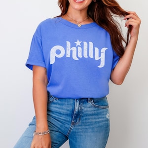 May include: A blue t-shirt with the word "philly" in white, stylized text and a star graphic. The shirt is paired with blue jeans. The model is wearing a bracelet and a necklace.