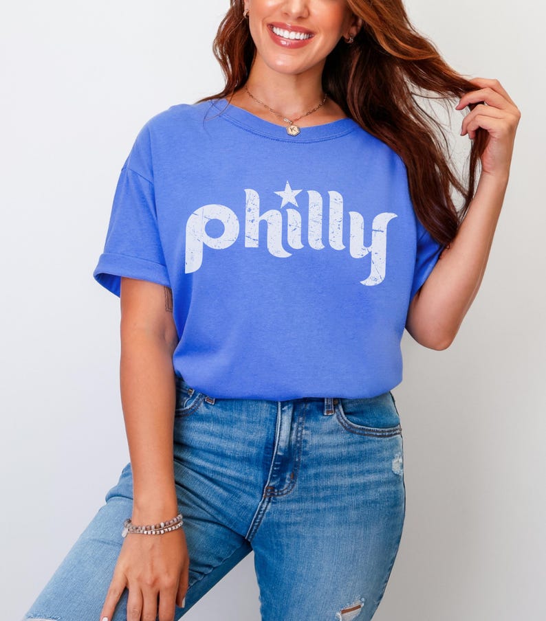 May include: A blue t-shirt with the word "philly" in white, stylized text and a star graphic. The shirt is paired with blue jeans. The model is wearing a bracelet and a necklace.