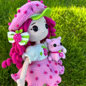 May include: A handmade crochet doll with a strawberry theme. The doll has a pink dress with red dots, a matching hat, and curly pink hair. It is holding a small pink cat. The doll is set against a green background.