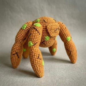 Puede incluir: Un juguete de araña hecho a mano, tejido a ganchillo, en tonos naranja y verde. La araña tiene ocho patas, un cuerpo redondeado y detalles verdes. Las patas y el cuerpo están detallados con costuras marrones.