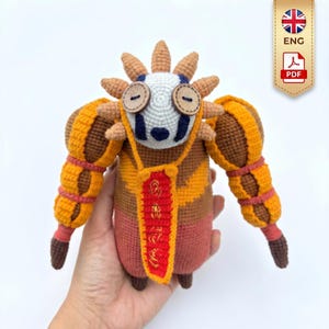 May include: A handmade crochet doll in shades of orange, brown, and pink. The doll has a sun-like headpiece, button eyes, and a red banner with gold text. The doll is held in a hand.