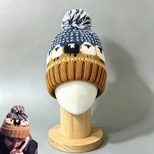Könnte beinhalten: Eine Strickmütze mit einem Muster aus weißen und schwarzen Schafen vor einem blau-beigen Hintergrund. Die Mütze hat einen gerippten beigen Umschlag am Saum und ist mit einem blau-weißen Bommel versehen. Das Design enthält ein kleines weißes Zickzackmuster.